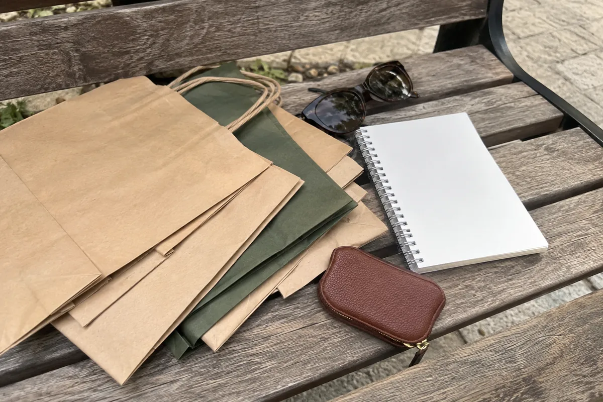 Folded shopping bags on a wooden bench beside a blank notepad, a pair of sunglasses and a small leather coin purse
